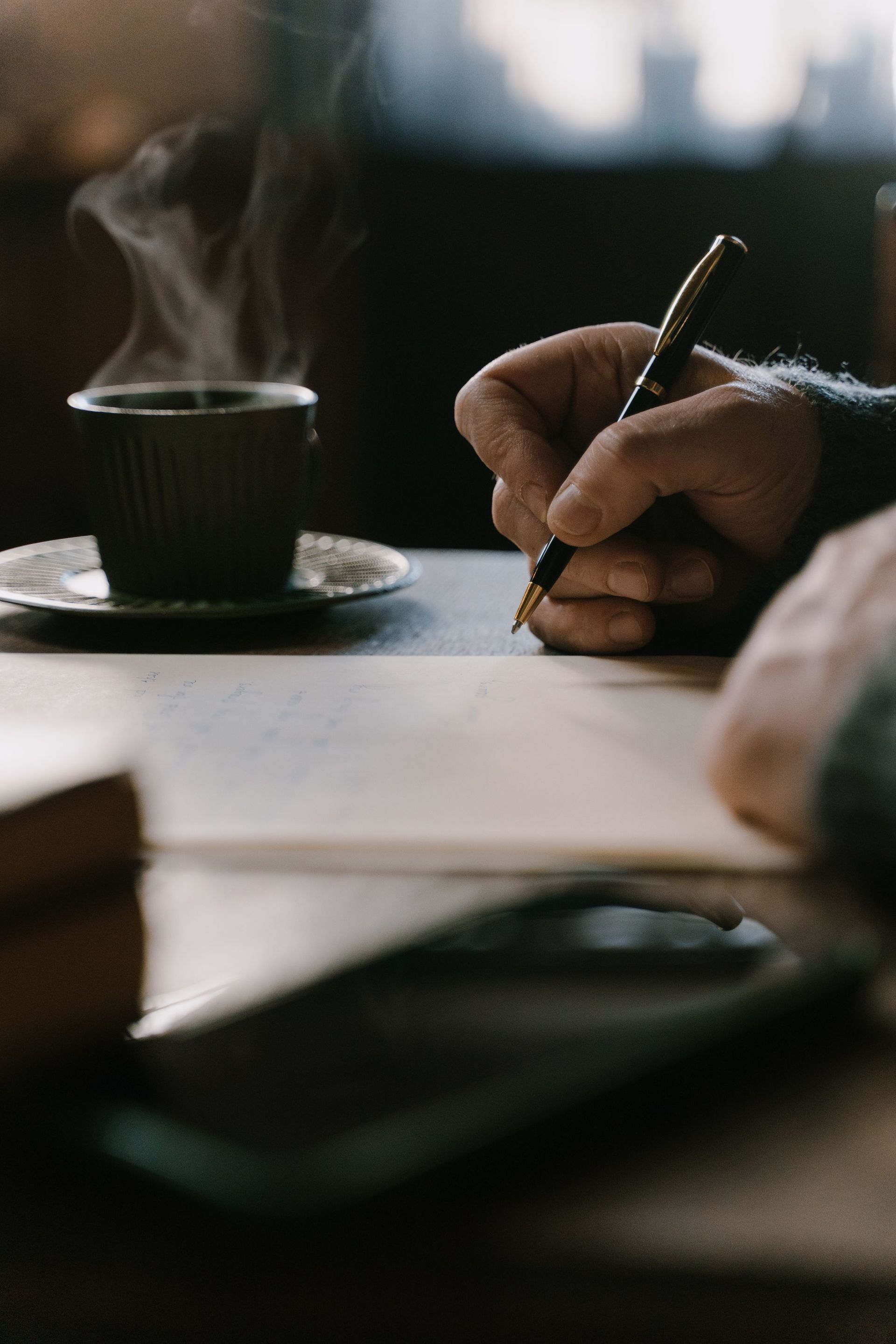 A warm scene of a person writing a note with a steaming coffee cup nearby.